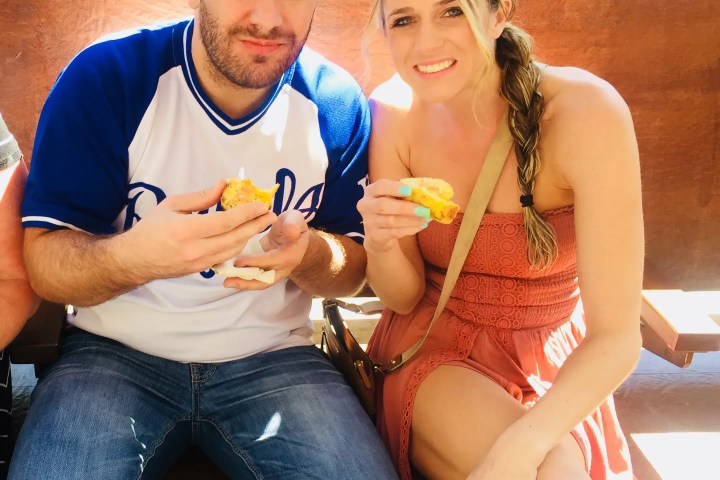 Man and woman sitting on bench eating sandwiches outdoors on a sunny day.