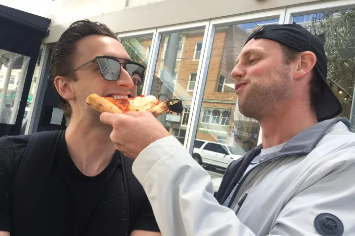 Two men, one feeding the other a slice of pizza, standing on a city street.