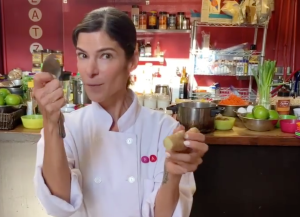 a woman chef in the foreground holding a spoon, with a kitchen in the background