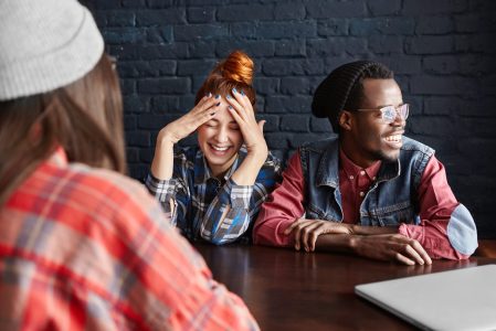 Pretty girl with ginger hair laughing out loud, holding hands on her head, keeping eyes closed during lively conversation with stylish African male in glasses and unrecognizable brunette female