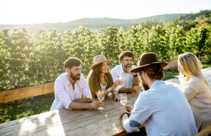 Group of a young people drinking wine and talking together while sitting at the dining table outdoors on the vineyard on a sunny evening