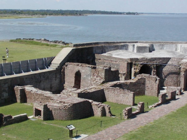 a wooden bench next to a body of water with Fort Sumter in the background