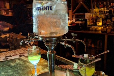 Absinthe fountain with green drinks on bar counter in dimly lit setting.