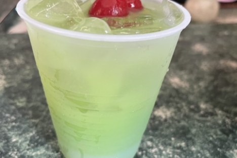 Green cocktail in a plastic cup with ice and a cherry garnish.