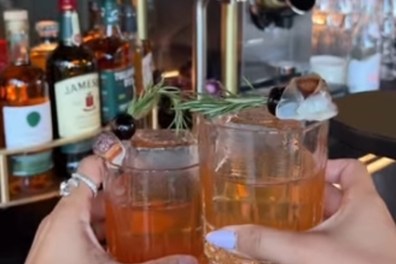 Two hands holding cocktails with rosemary and olives at a bar.
