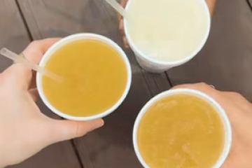 Three hands holding frozen drinks in white cups with straws over a wooden surface.