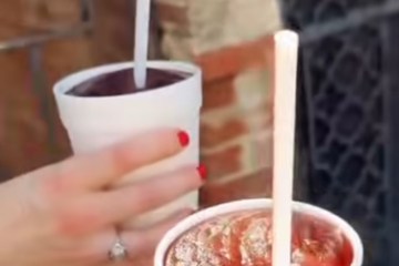 Two hands holding slush drinks in white cups with straws outdoors.