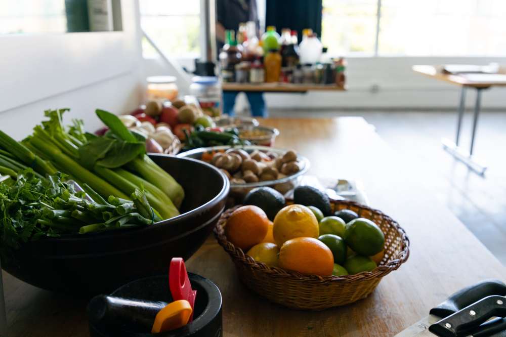Table with bowls of fruits, vegetables, and cooking ingredients in a bright kitchen.