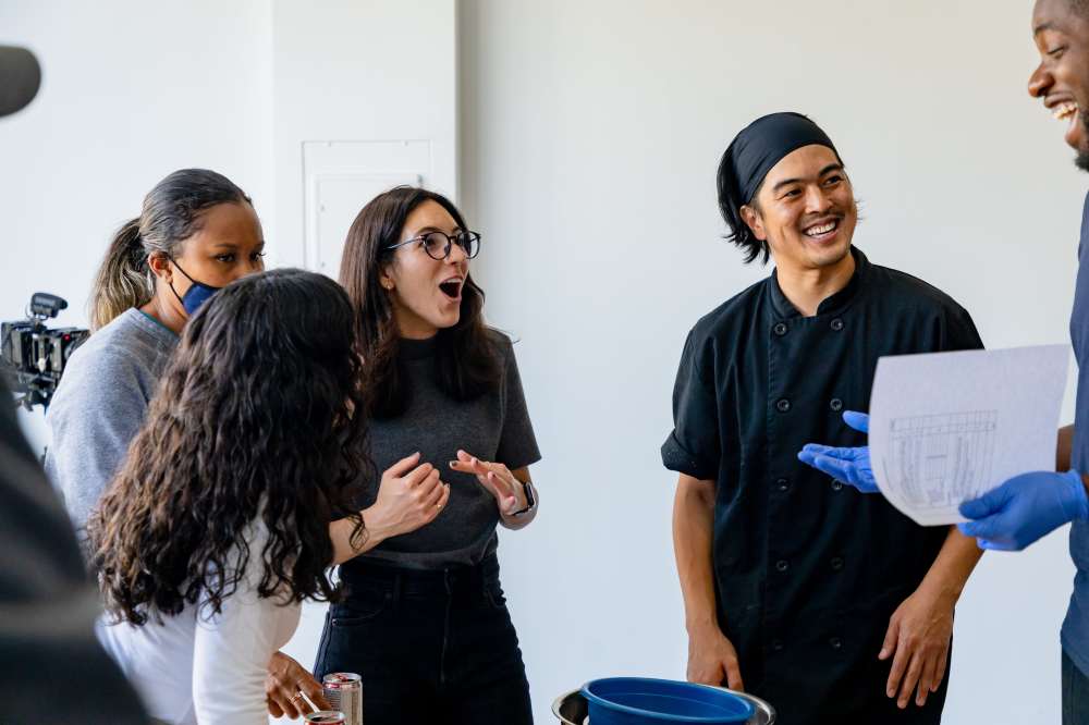 Group of people laughing and talking, one holding paper, in a casual setting.