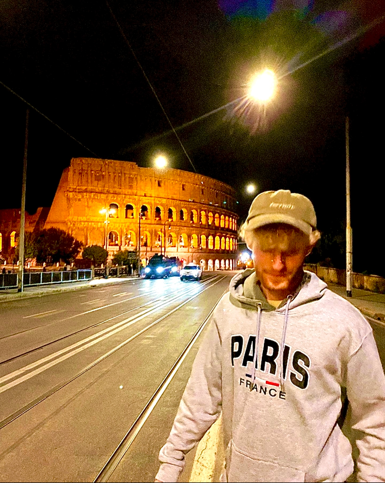Person in hoodie stands by the Colosseum at night with street lights illuminating the scene.