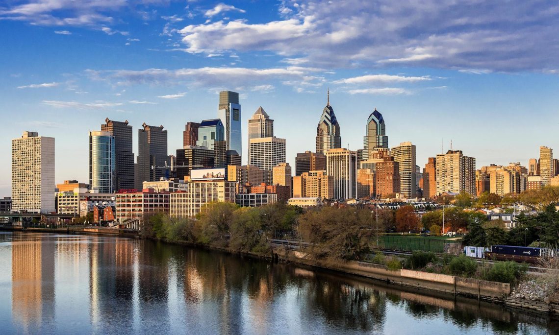 Philadelphia skyline with skyscrapers reflecting in a river under a blue sky.
