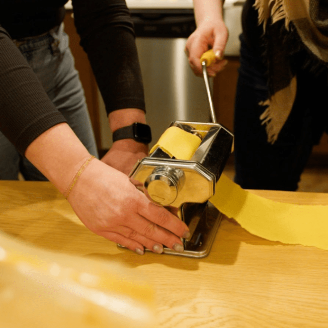 Two people using a pasta machine to roll out fresh pasta on a wooden table.