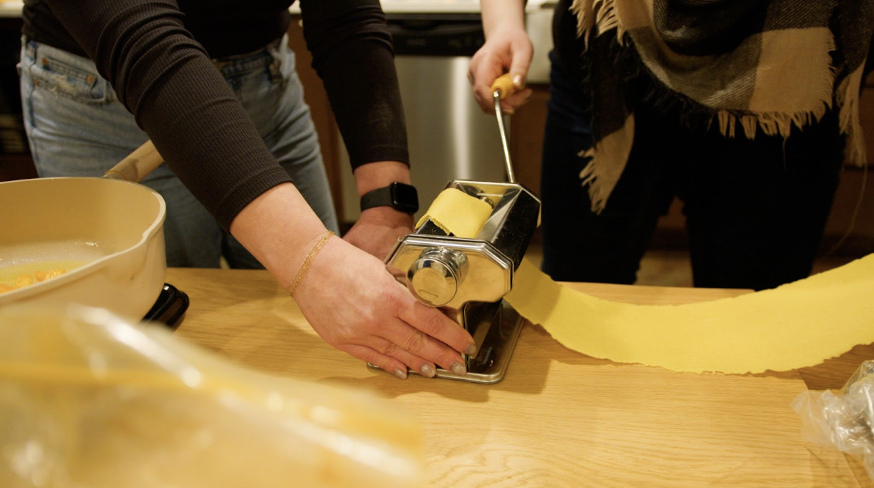 Two people using a pasta machine to roll out fresh pasta on a wooden table.
