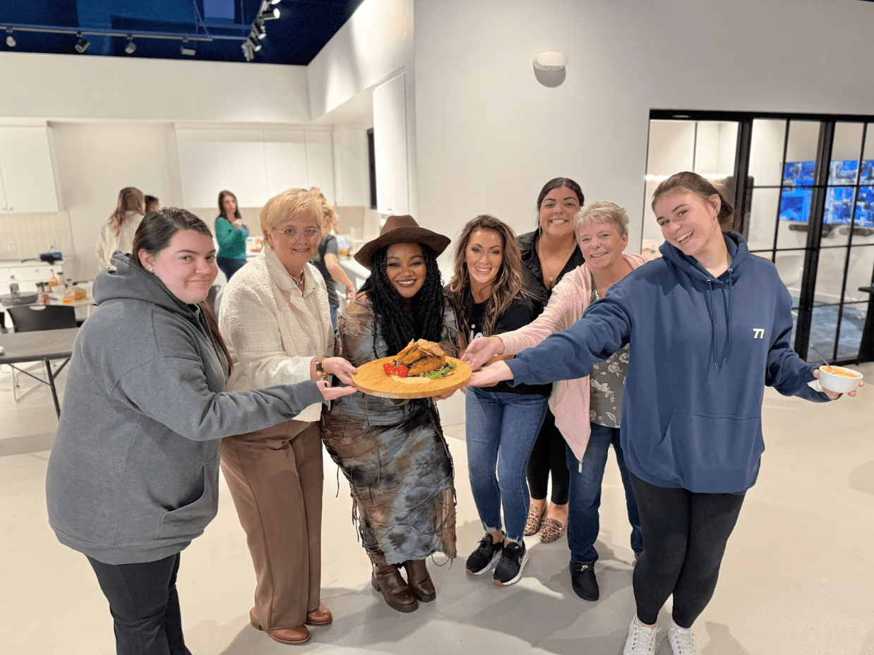 Group of seven people smiling, holding a plate of food in an indoor setting.