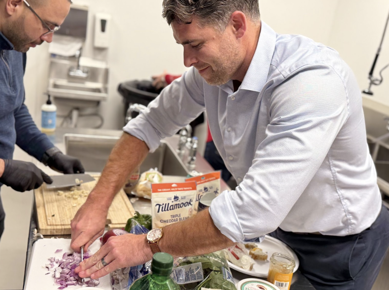 Two men in a kitchen chopping onions, surrounded by ingredients and Tillamook cheddar cheese.