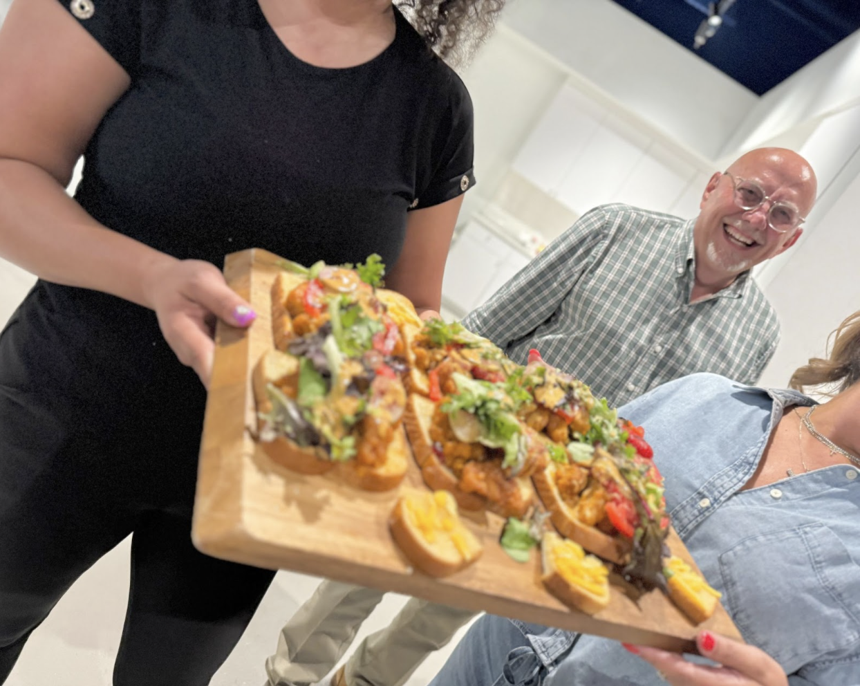 People holding a wooden board with loaded sandwiches, one man smiling in the background.