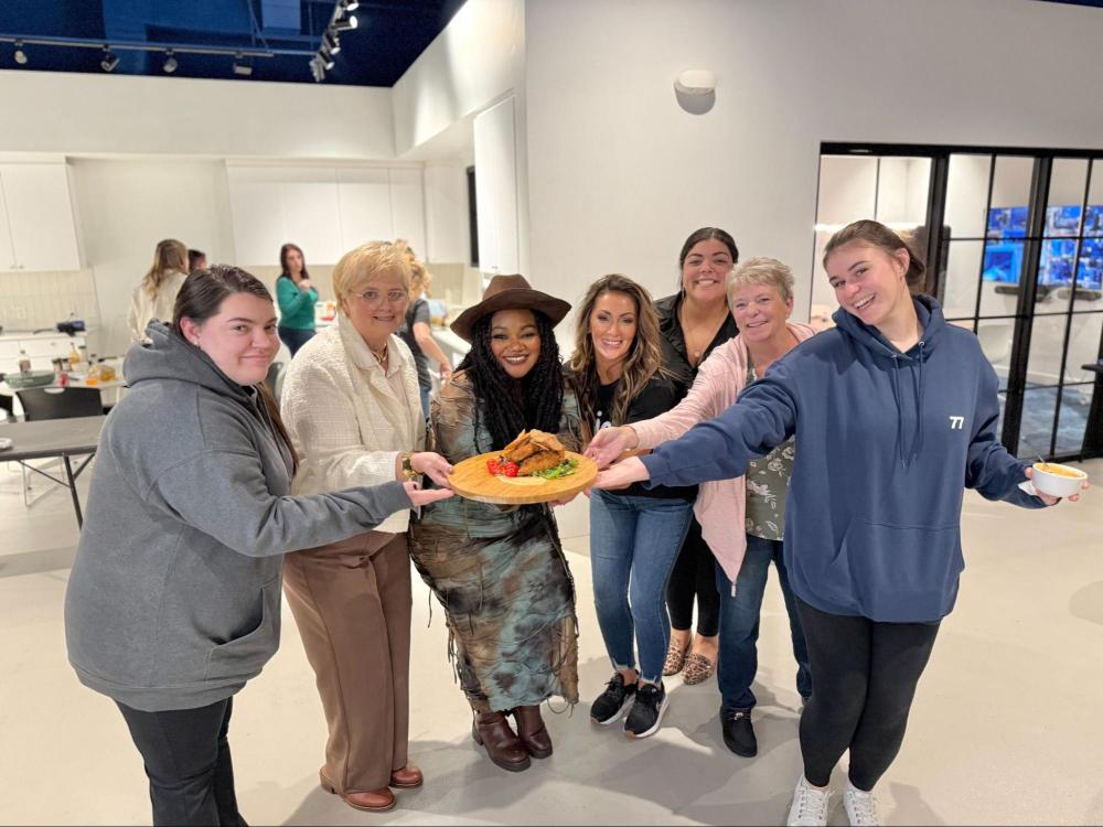 Six women in a kitchen posing with a wood tray of food, smiling towards the camera.