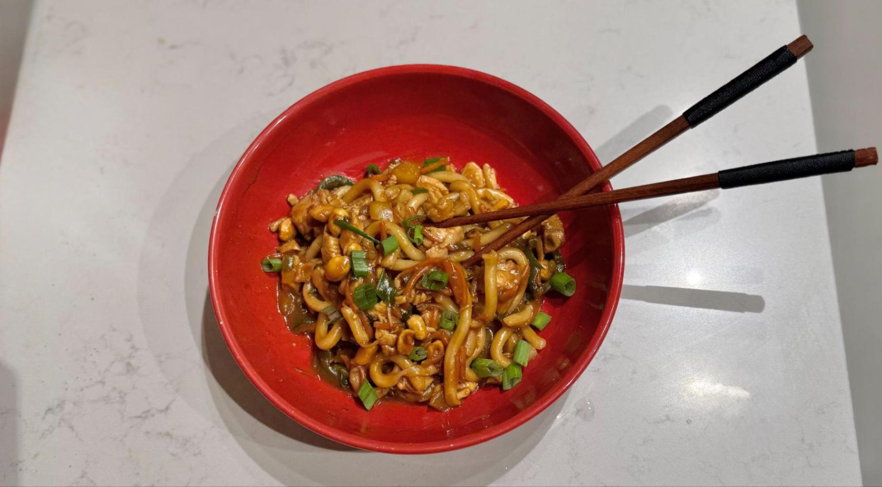 Red bowl with stir-fried noodles, vegetables, and chopsticks on a white table.