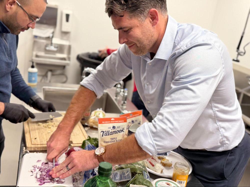 Two men chopping vegetables on a kitchen counter with cheese packaging visible.