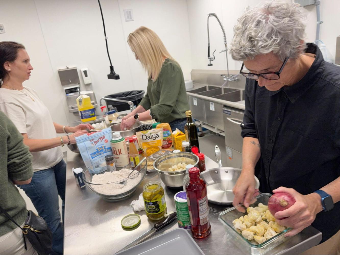 Three people cooking with various ingredients on a stainless steel counter in a kitchen.