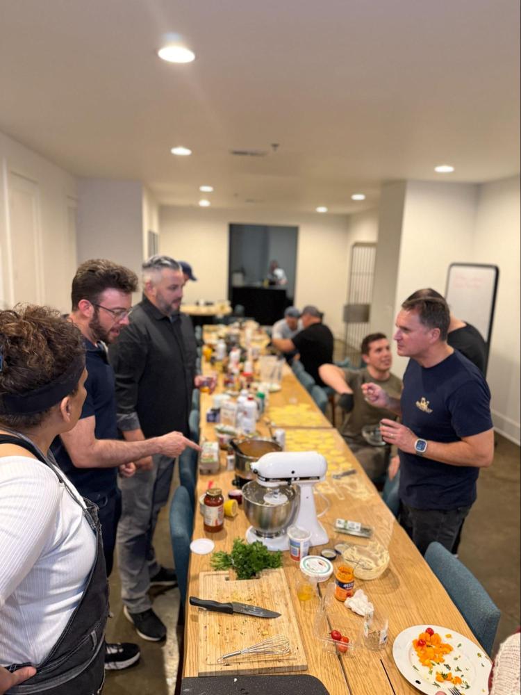 Group of people gathered around a long kitchen table with cooking equipment and ingredients.