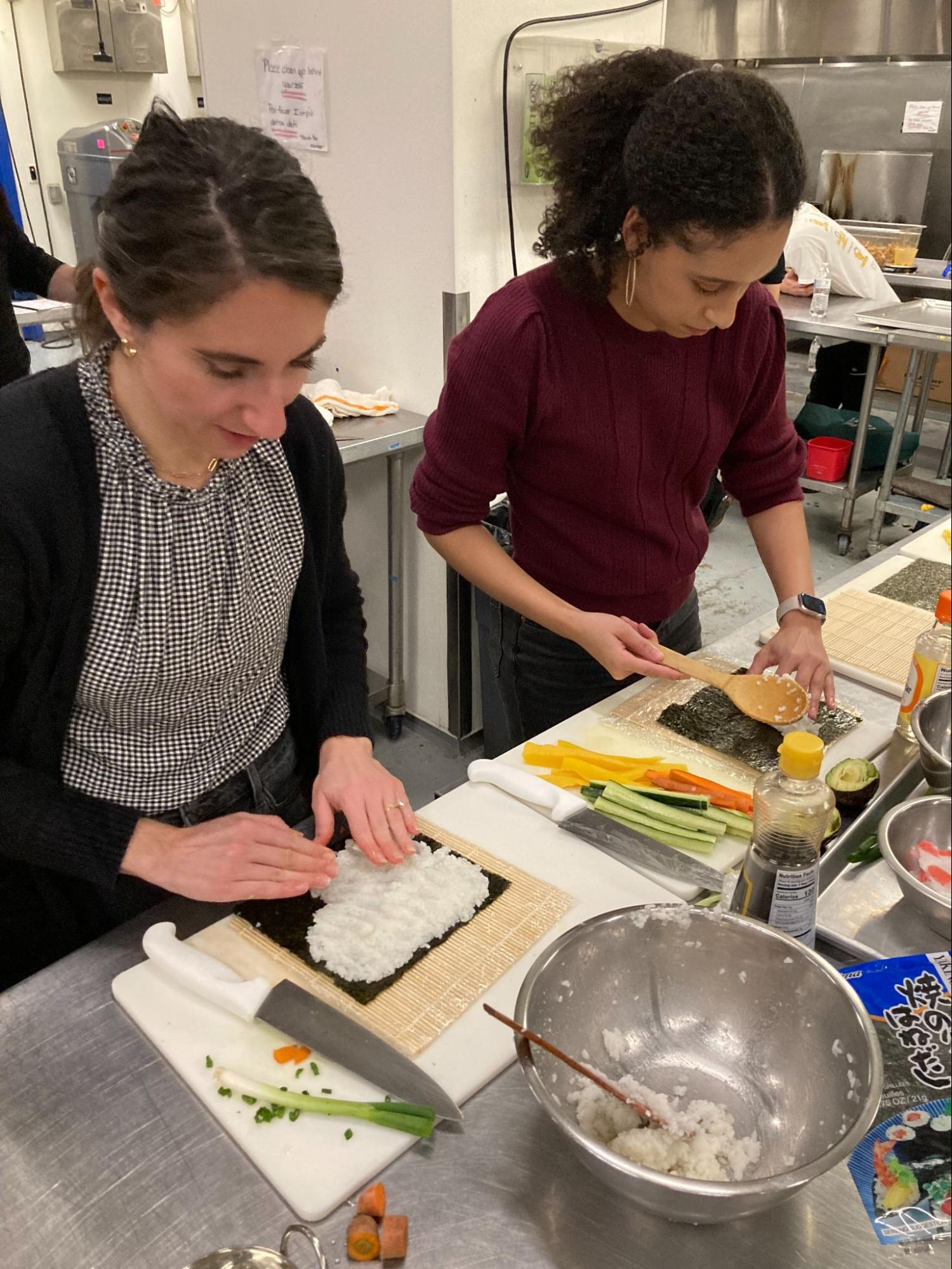 Two women preparing sushi rolls with rice and seaweed in a kitchen setting.