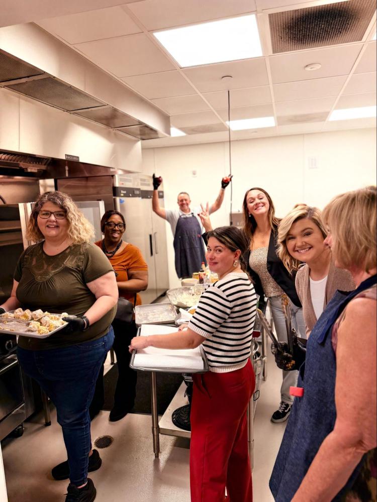 Group of people smiling in a commercial kitchen, holding trays and wearing casual clothes.