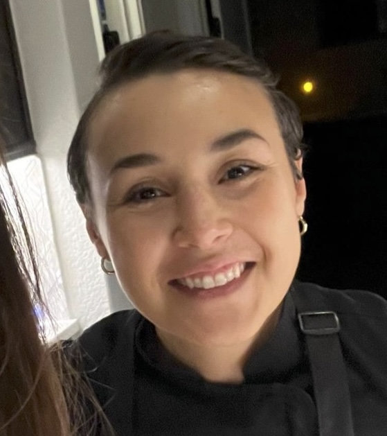 Person smiling, wearing a black outfit with short hair and hoop earrings.