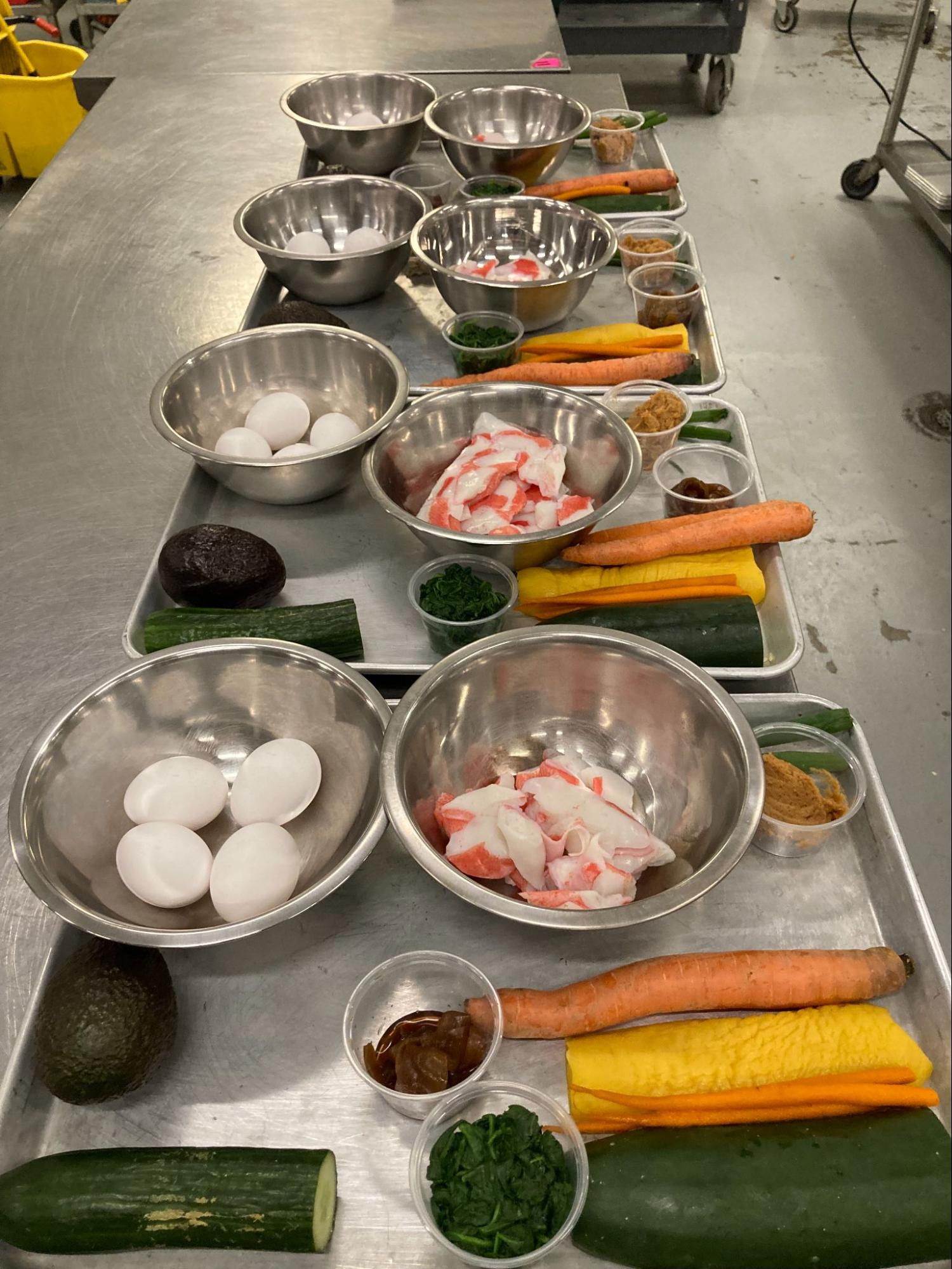 Metal bowls with eggs, imitation crab, carrots, cucumbers, and small ingredient cups on a prep table.