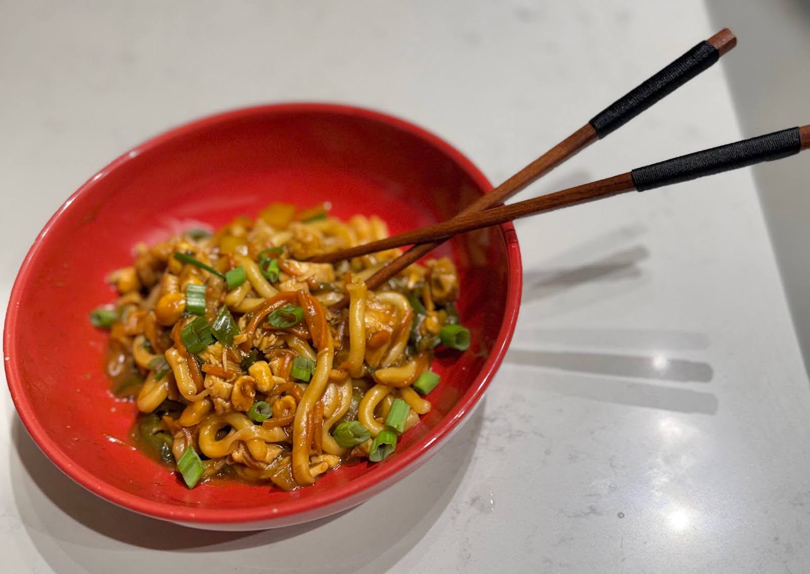 Noodles with vegetables and chopsticks in a red bowl on a countertop.