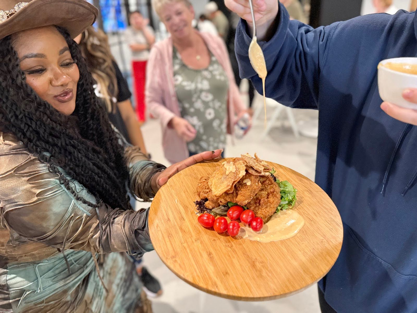 Person holding a wooden platter with crispy chicken, cherry tomatoes, greens, as sauce is poured on the side.
