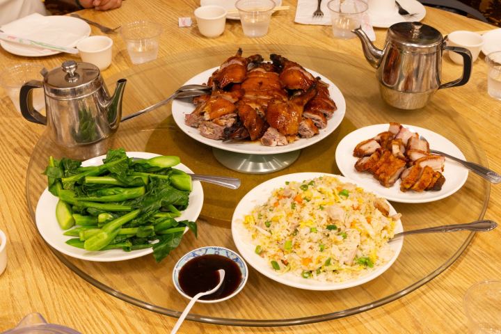 Chinese meal with roast duck, stir-fried greens, fried rice, and tea pots on a round table.