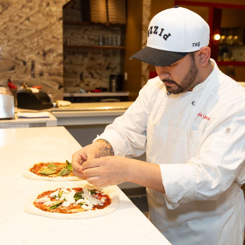 Chef prepares pizzas with toppings in a kitchen setting.
