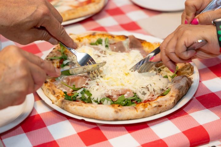 Hands with forks near a pizza on a red plaid tablecloth.