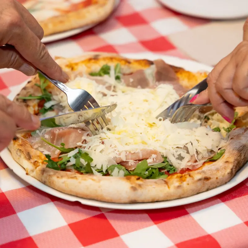 Hands with forks near a pizza on a red plaid tablecloth.