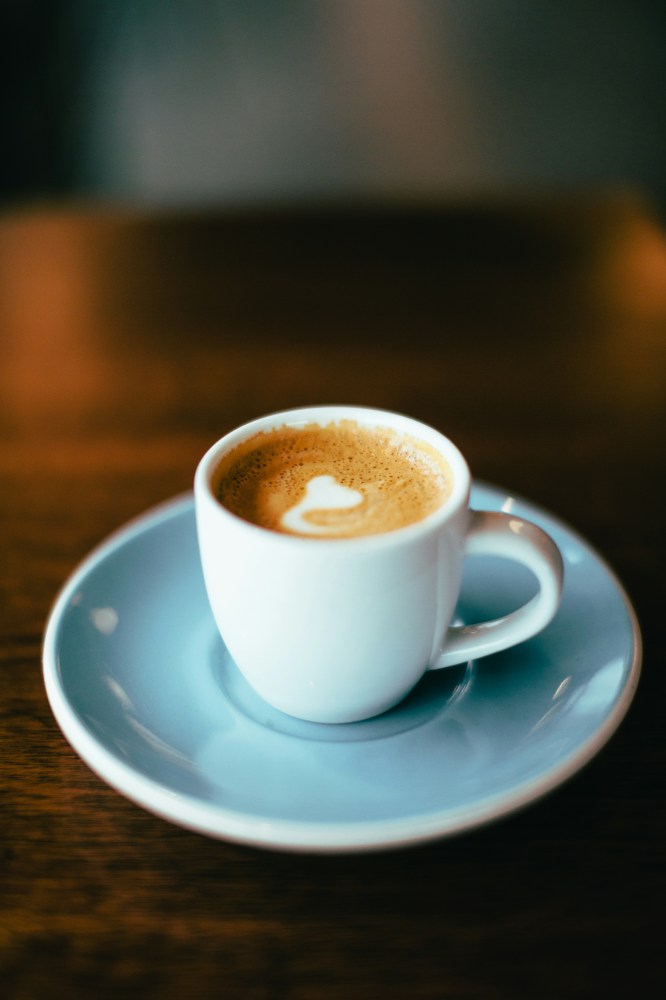 White cup of coffee with foam heart on light blue saucer, blurred background.